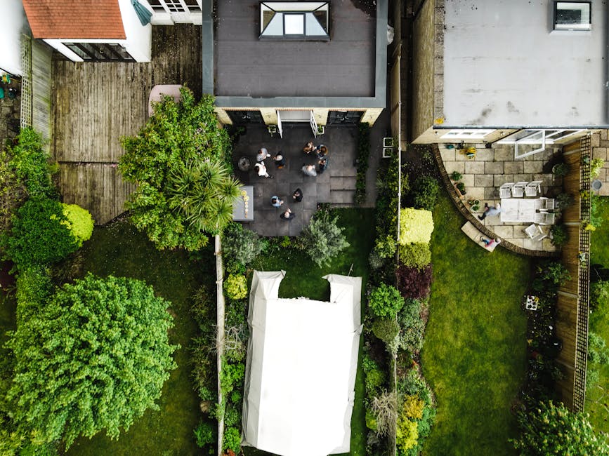 An aerial view of a residential garden during daytime, showcasing a diverse landscape with well-maintained lawn areas, lush green trees, and flower beds along the borders. In the center, a paved patio