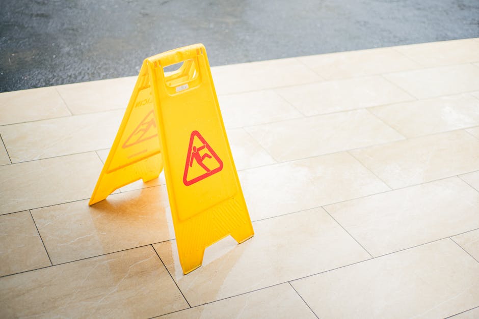 A bright yellow, A-frame wet floor warning sign with a red triangular caution symbol featuring a person slipping, placed on a beige tiled outdoor patio area. The tiles have a smooth, polished surface 