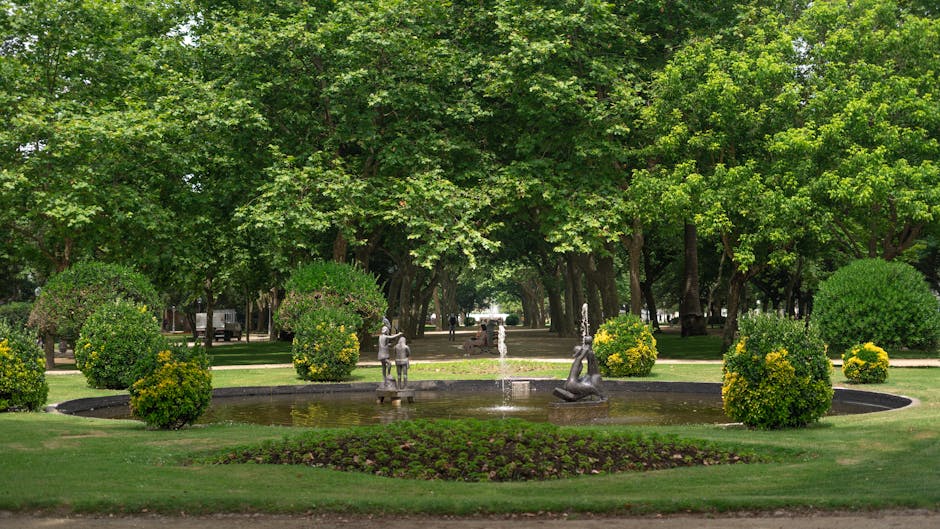 A serene garden scene in Gladstone Park featuring a lush, well-maintained lawn with neatly trimmed grass, bordered by dense greenery including bushes and trees with fresh green leaves. In the foregrou